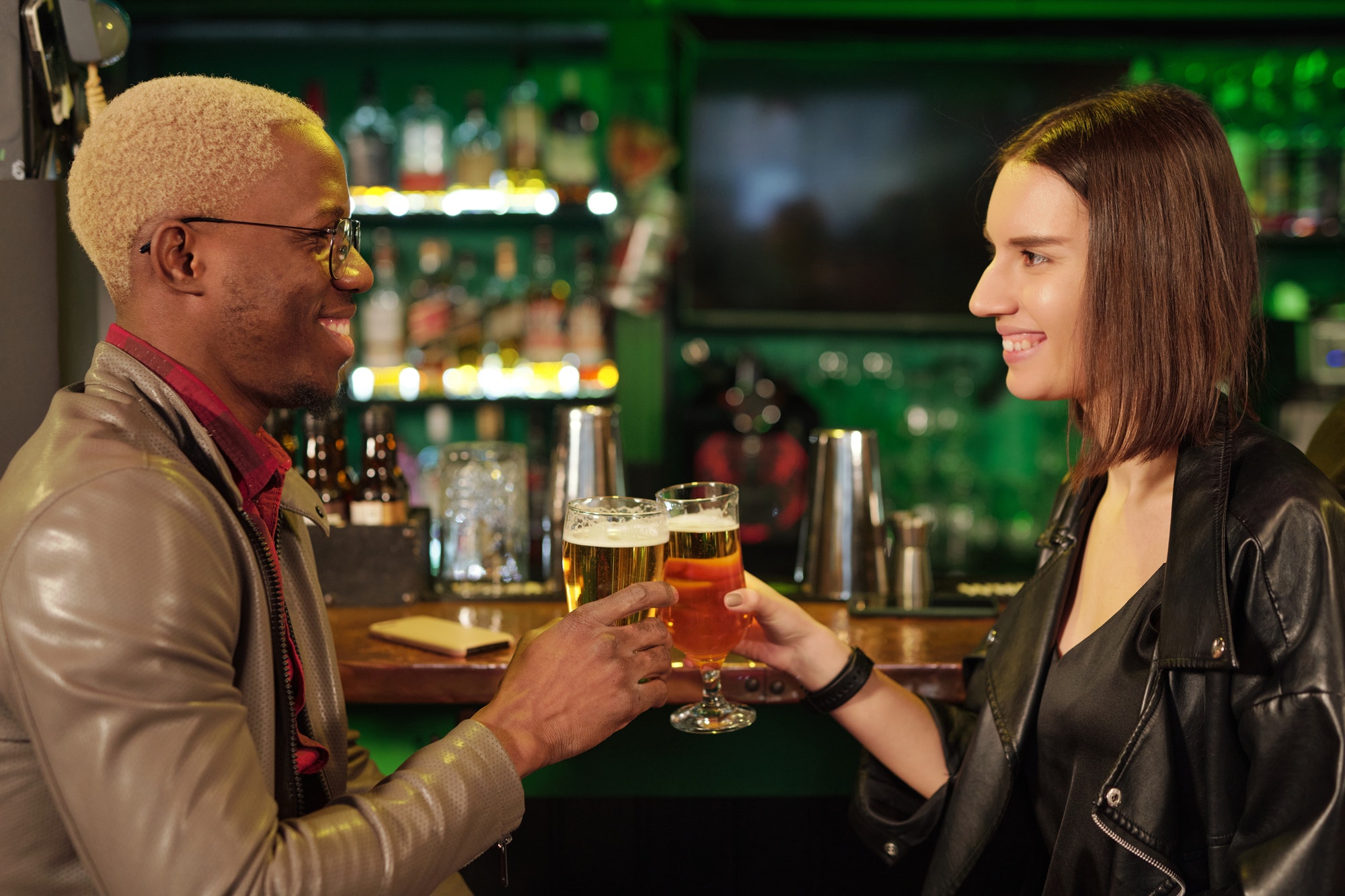 Happy young African man and his girlfriend clinking with glasses of beer in bar